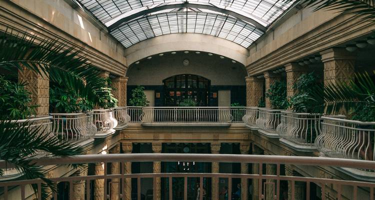 Grand hall intérieur avec toit en verre teinté, plantes de palmiers et balustrades ornées, ressemblant à un spa historique.