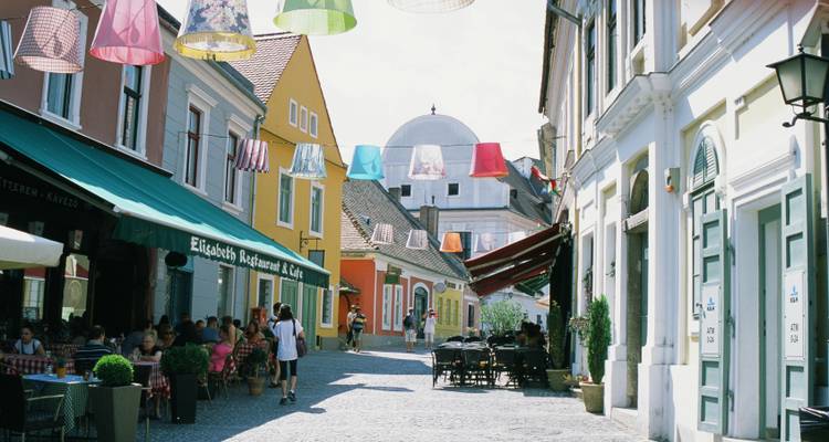 Rue piétonne colorée à Szentendre bordée de cafés et de lampadaires suspendus par un jour ensoleillé.