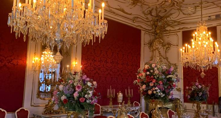 Lujoso comedor rococó en rojo y oro adornado con lámparas de cristal y arreglos florales.