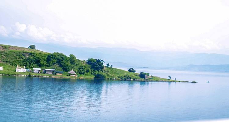 Helder, rustig gedeelte van het Kivu-meer met een klein grazig schiereiland en verre bergen.