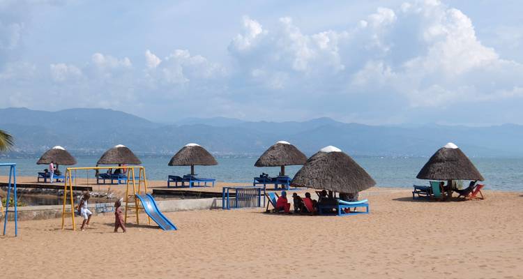 Sandy lakeside beach with blue lounge platforms, thatched shade huts, a small playground and distant hazy mountains across the water.