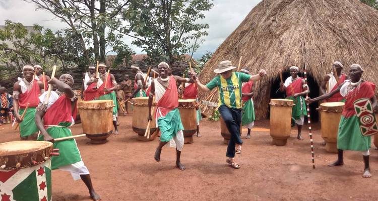 Traditional drummers in colourful attire perform while a tourist joins in front of a thatched hut village setting.