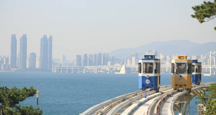 Zwei farbige Küsteneinschienenbahn-Wagen fahren über erhöhte Gleise mit Busan-Skyline und Meereshintergrund.