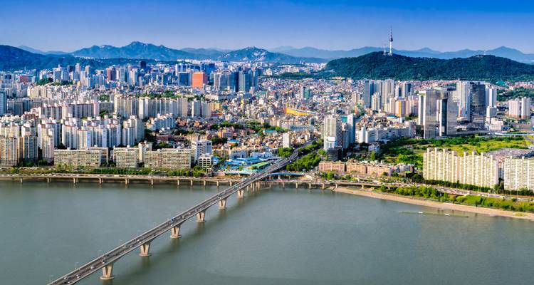 Panoramischer Luftaufnahme der Seouler Skyline entlang des Han-Flusses mit langer Brücke und dem Namsan Tower auf einem bewaldeten Hügel.