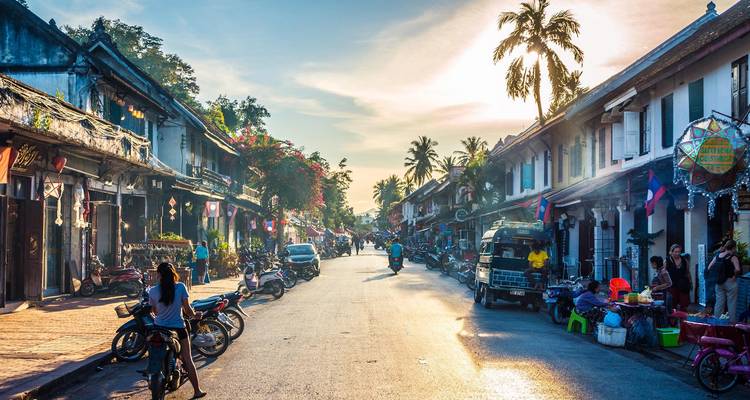 Rue coloniale ensoleillée bordée de boutiques et de motocyclettes à Luang Prabang à l'approche de la soirée.