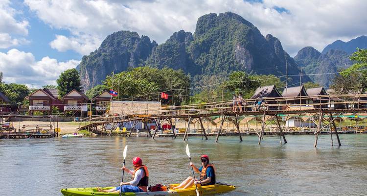 Deux kayakistes pagaient sur une rivière calme sous des montagnes de calcaire imposantes et un pont de bambou.