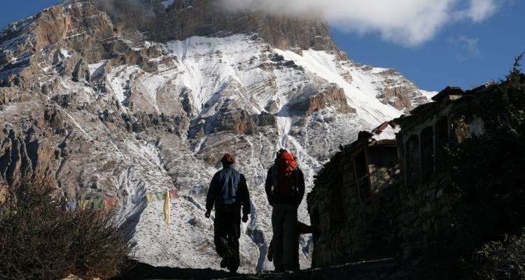 Twee trekkerslopers lopen over een rotsig pad onder een imposante, met sneeuw bedekte Himalayatop die in het ochtendlicht baadt.