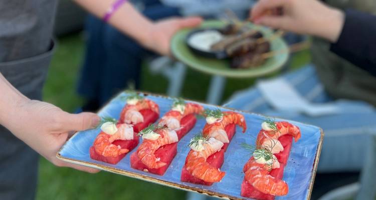 El camarero presenta un plato de camarones rojo brillante en canapés mientras otra persona sostiene pinchos en el fondo.