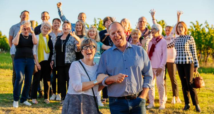 Grupo grande y alegre posando en un viñedo al atardecer con dos personas riendo en primer plano.