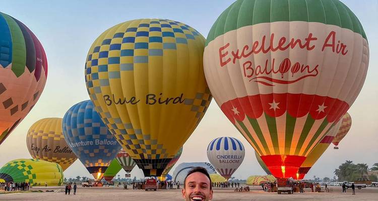 Un voyageur souriant se tient devant une rangée de montgolfières colorées prêtes pour le décollage à l'aube.