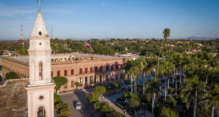 Vue aérienne d'un bâtiment colonial avec un clocher blanc élancé et une place entourée de palmiers sous un ciel bleu dégagé