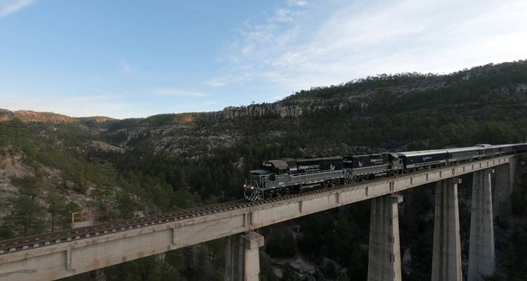 Plan large du train Chepe Express traversant un pont en béton imposant à travers un terrain de canyon accidenté