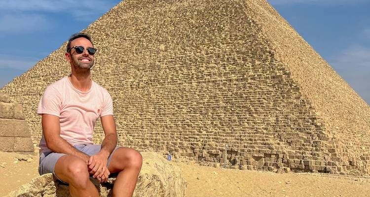 Un voyageur sourit assis sur un bloc de pierre avec la Grande Pyramide se dressant derrière.