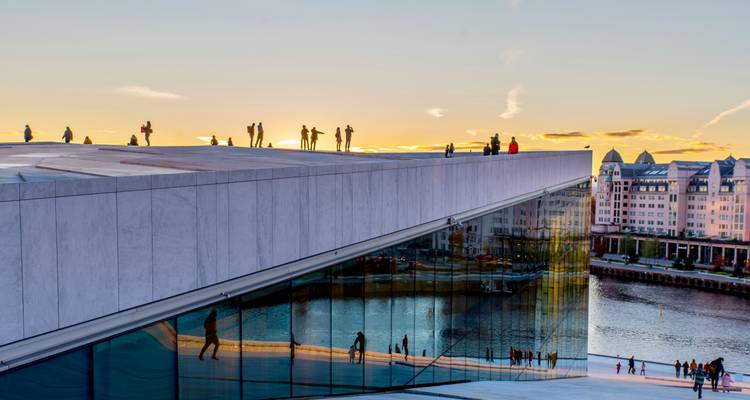 People walking on the sloped roof of Oslo Opera House at sunset with reflections in the glass facade.