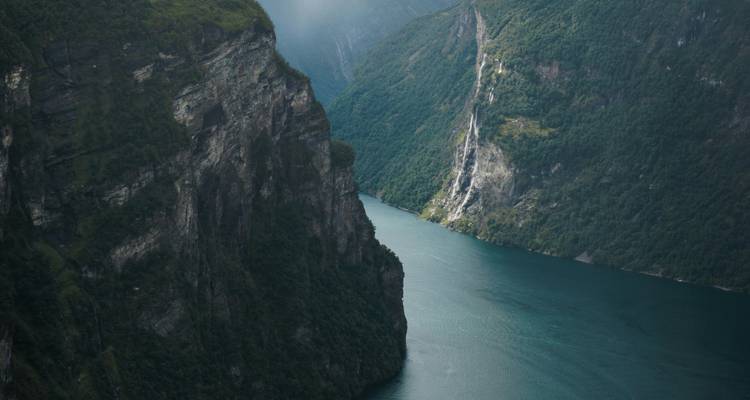 Steep dark cliffs plunge into a deep blue fjord under misty light.