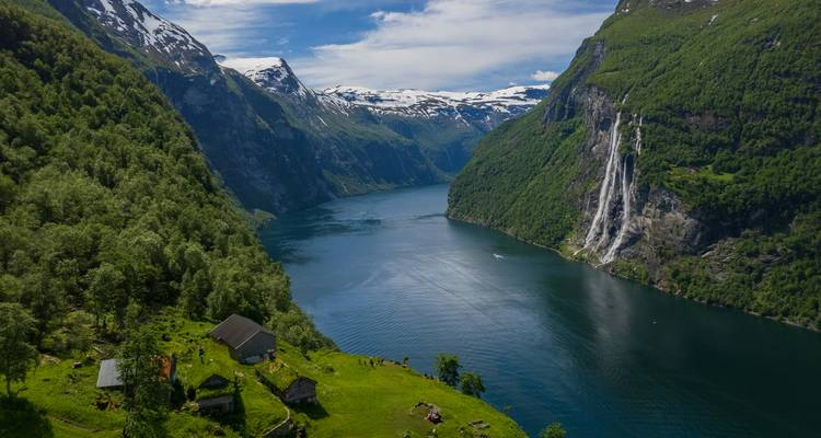 Panoramic aerial of a fjord with waterfalls, snow-capped peaks and green pastures.