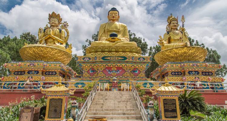 Trois grandes statues dorées de Bouddha assis sur une plateforme richement décorée sous un ciel partiellement nuageux.