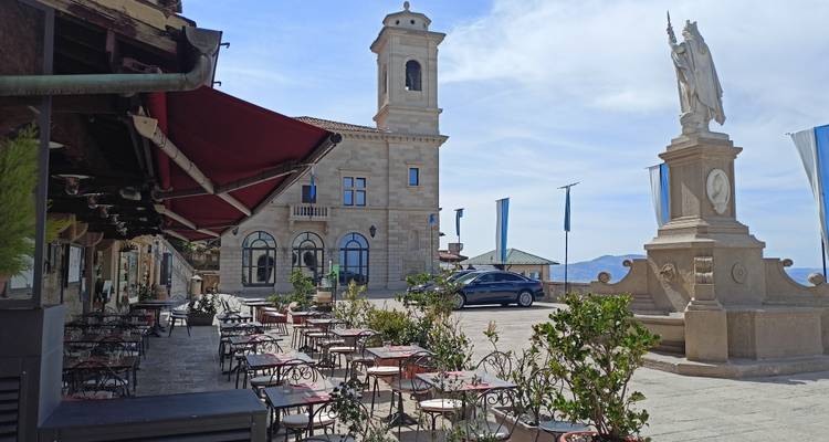 Terrace café with empty tables beside a stone bell tower and statue on a sunny hilltop plaza.