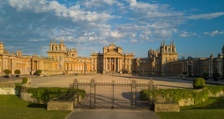 Grand wrought-iron gates framing full facade of Blenheim Palace bathed in warm early light.