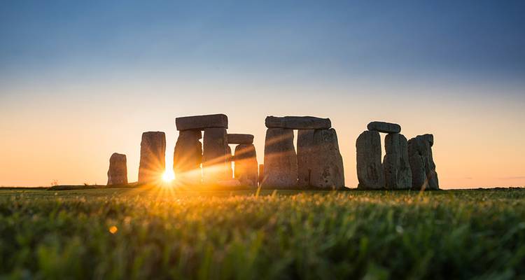 Iconic Stonehenge stone circle silhouetted as sunrise rays burst through the monoliths over dewy grass.