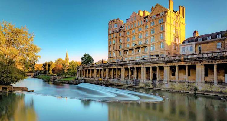 Historic architecture of Pulteney Bridge and weir reflected in smooth river water at golden hour in Bath.