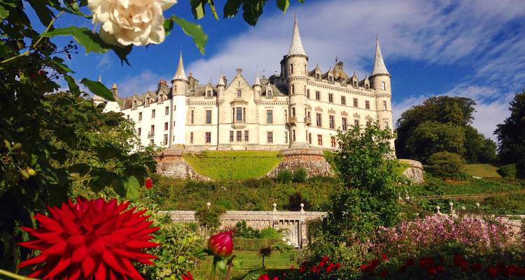 Fairytale Dunrobin Castle framed by vibrant red and white flowers under a bright blue sky.