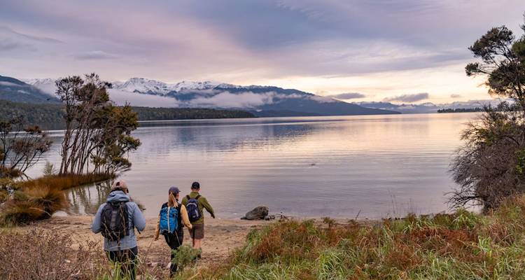 Les randonneurs s'approchent d'un lac paisible à l'aube avec des montagnes enneigées et de bas nuages au-delà.