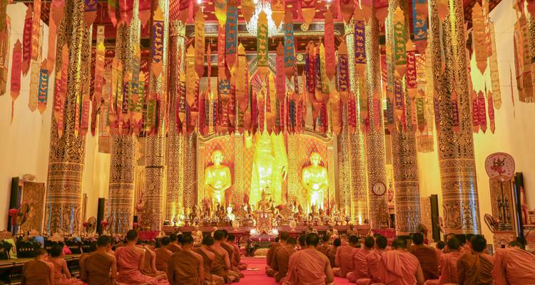 Intérieur d'un temple bouddhiste baigné de lumière dorée où des dizaines de moines sont assis devant des statues ornées lors d'une cérémonie.