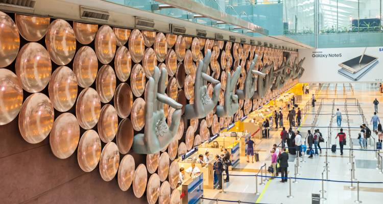 Amplia sala de terminales del Aeropuerto de Delhi adornada con un gigantesco muro de arte con mudra de mano de cobre y viajeros ocupados abajo.