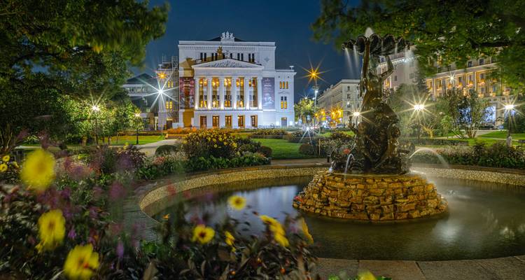 Vue nocturne de l'Opéra National de Riga illuminé derrière une fontaine et des parterres de fleurs colorés.