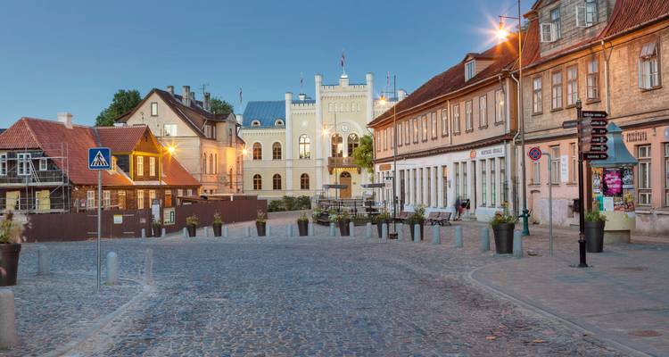 Scène de soirée doucement éclairée d'une place pavée de galets bordée de bâtiments historiques et de lanternes.