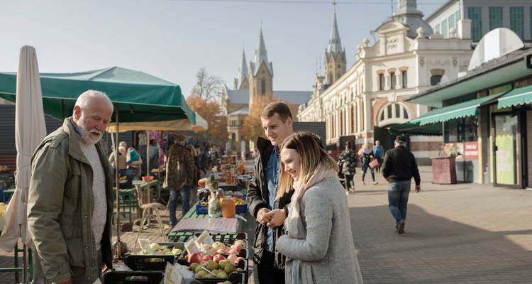 Mercado callejero al aire libre donde los locales curiosean frutas y verduras con el telón de fondo de edificios históricos.