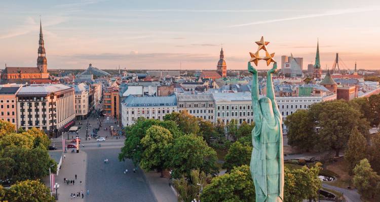 Vista aérea al atardecer sobre Riga con el Monumento a la Libertad en primer plano dominando el horizonte de la ciudad vieja.
