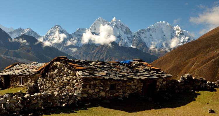 Refuge de berger en pierre avec toiture en ardoise dans un pré alpin élevé encadré par des pics enneigés imposants.