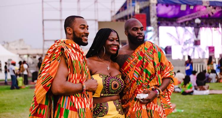 Trois jeunes adultes vêtus de tissu Kente aux couleurs éclatantes posent ensemble lors d'un festival culturel en plein air avec une scène en arrière-plan.
