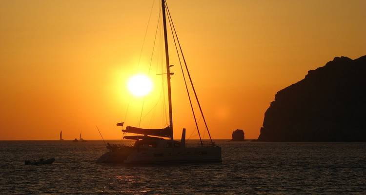 Silhouette d'un catamaran ancré face à un coucher de soleil orange vif avec des voiles lointaines et une côte accidentée.