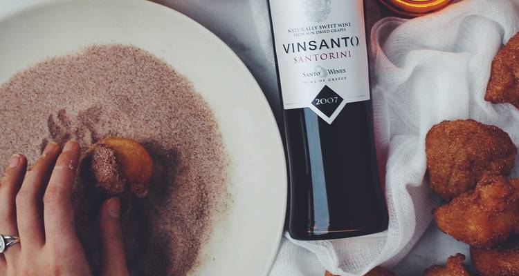 Une main enrobe un chaud feuilleté de sucre à la cannelle à côté d'une bouteille de vin de dessert de Santorin sur une table.
