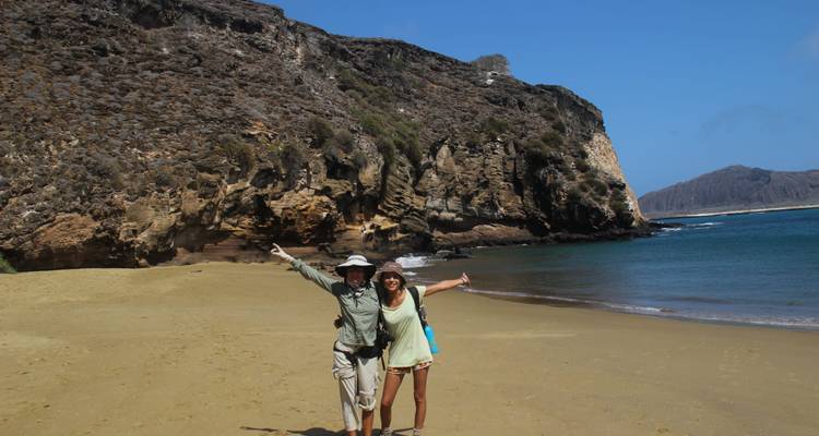 Pareja feliz con los brazos extendidos de pie en una playa de arena vacía respaldada por acantilados escarpados y mar azul.
