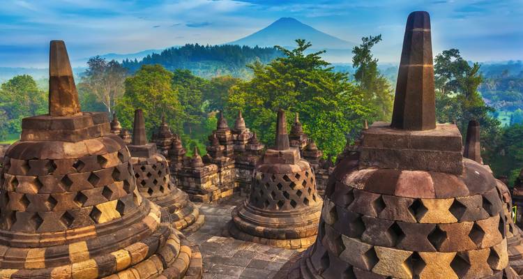 Stenen stupas van Borobudur als silhouet tegen een mistig bos en vulkaan in de verte bij zonsopgang.