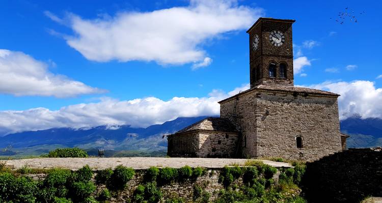 La torre de reloj de piedra del Castillo de Gjirokastër se alza contra un cielo azul brillante y montañas distantes.