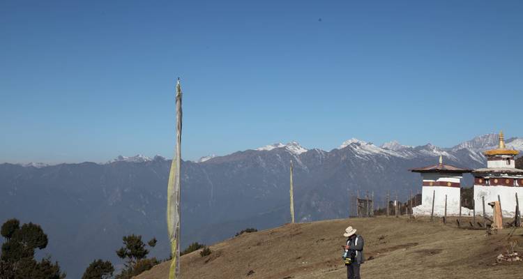 Un excursionista está de pie junto a un pequeño chorten y banderas de oración en un alto paso de montaña bajo un cielo azul despejado.