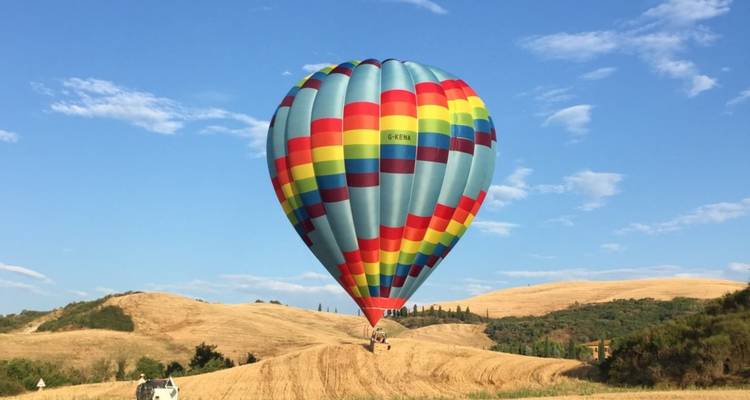 Un colorido globo aerostático flota justo encima de los dorados campos toscanos bajo un cielo azul brillante.