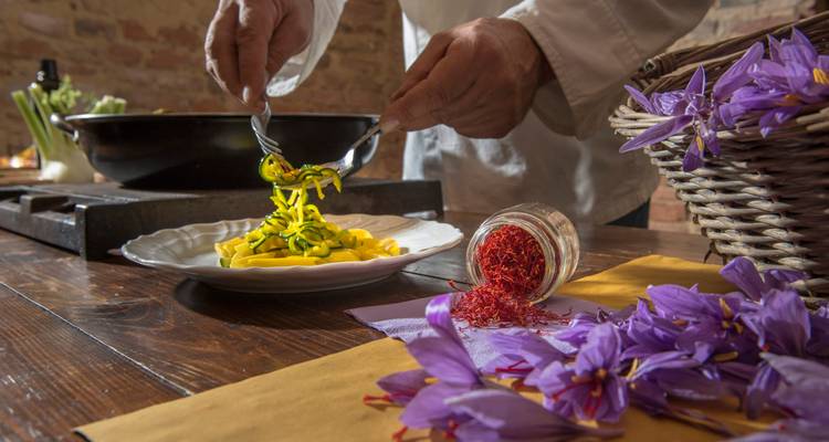Un chef sirve pasta fresca con azafrán en una cocina rústica, con flores púrpura vívidas y especias cerca.