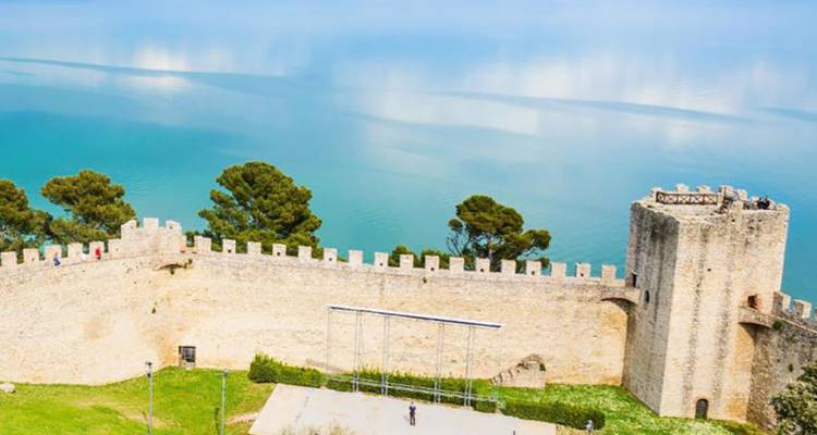 Las murallas de un castillo de piedra se alzan sobre un vasto lago turquesa rodeado de árboles frondosos.