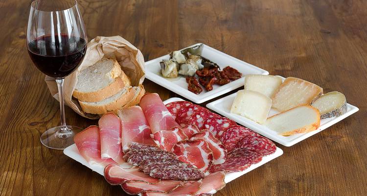 Una mesa de madera con embutidos curados, quesos, pan y una copa de vino tinto.