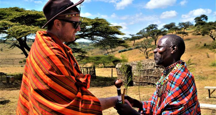 Anciano maasai y viajero masculino vestidos con ropa tradicional estrechándose las manos en la sabana cubierta de pasto.