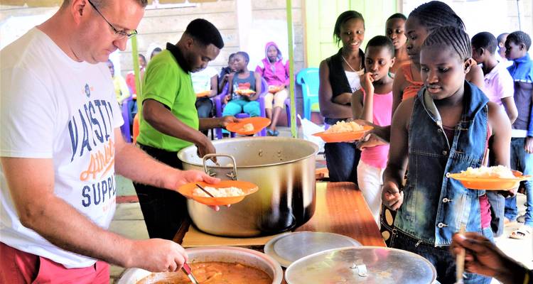 Un voluntario sirviendo comida de grandes ollas a niños en un área de comedor comunitario.