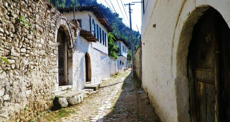 Une ruelle pavée étroite bordée de pierre et de maisons ottomanes chaulées sous des collines verdoyantes.