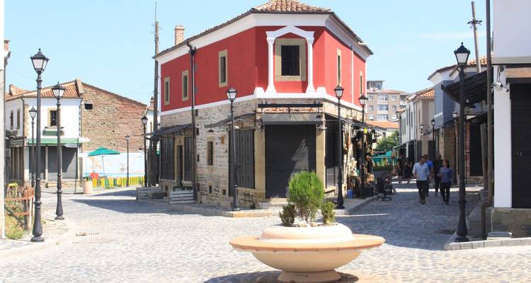 Une place de bazar ottoman restaurée avec un bâtiment rouge frappant, des rues en pierre, des réverbères et un petit groupe de piétons.
