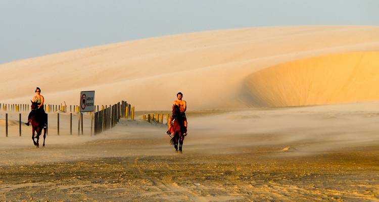 Two horse riders crossing a windy sandscape at dusk with rolling dunes behind